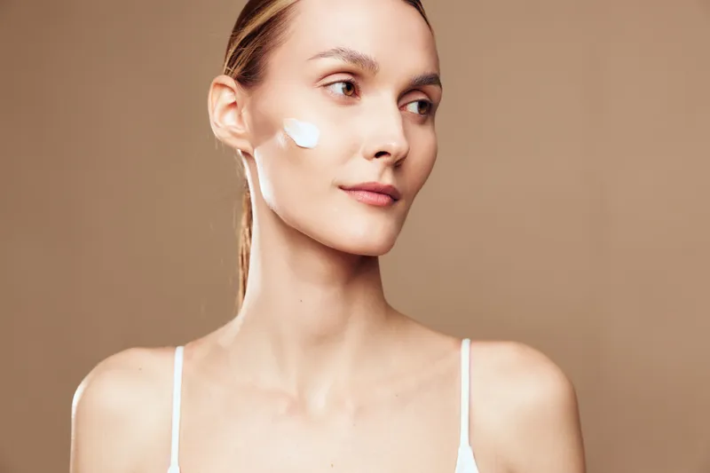 A young woman with fair skin applies white cream on her cheek, looking sideways against a neutral beige background. Concept for skincare, beauty, and self-care routine.