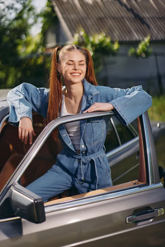 Cheerful young woman in casual denim outfit smiling from car window on a sunny day, reflecting joy and freedom in nature.
