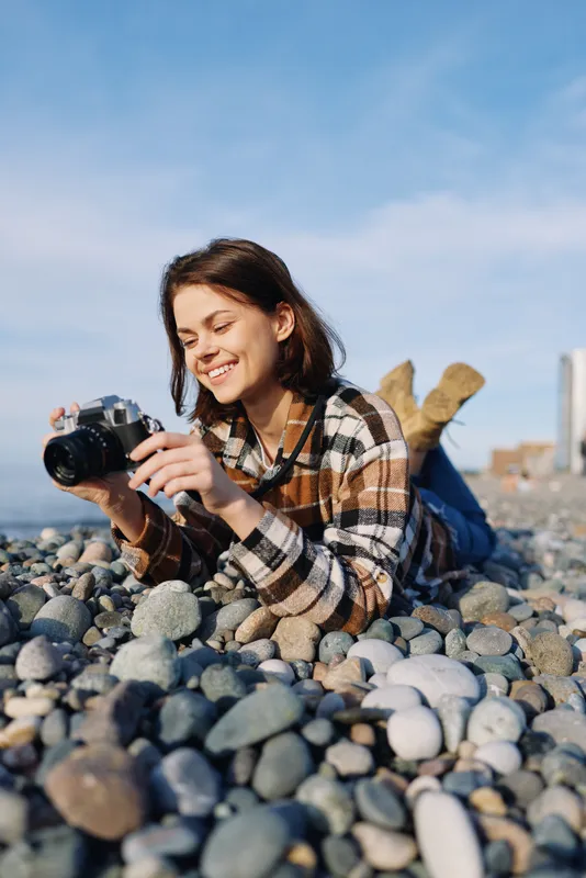 Female photographer lying on a pebble beach, smiling while adjusting a camera, outdoor hobby shot with natural light, casual plaid shirt and bright sky behind.