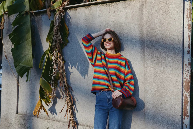 Woman portrait with sunglasses and colorful sweater leaning against wall, smiling in casual jeans and crossbody bag, sunny outdoor fashion scene with relaxed urban vibe.