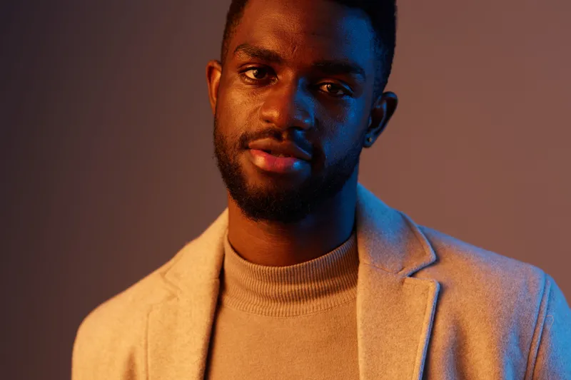 Portrait of a young Black man in a stylish beige blazer, expressing confidence and sophistication against a soft blue and orange gradient background