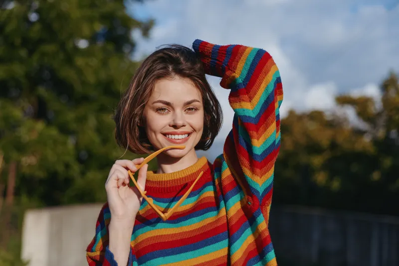 Smiling woman portrait in colorful sweater outdoors, young woman holding sunglasses, casual and happy expression in a sunny park, cozy knit, short brunette hair and relaxed mood.
