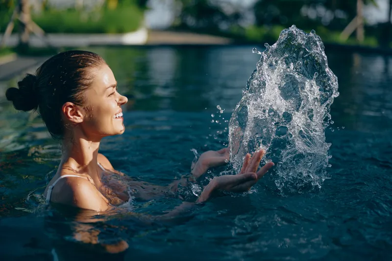 Smiling woman in a pool splashing water with hands, enjoying outdoor leisure activity. Bright natural light highlights joyful expression and relaxed lifestyle moment.