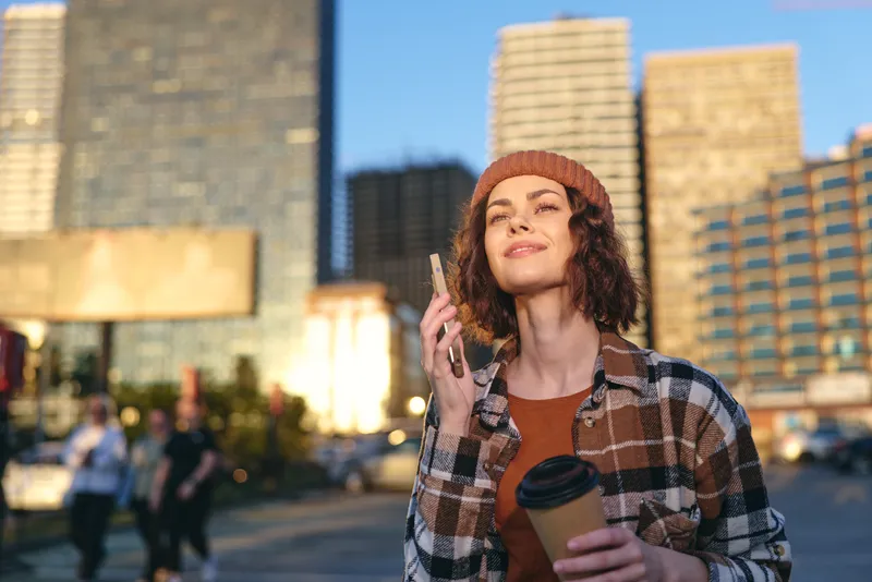Woman with coffee and phone in city plaza, smile lit by golden hour glow and sunlight, beanie and plaid jacket convey authenticity and candid lifestyle, mindful living and emotional storytelling.