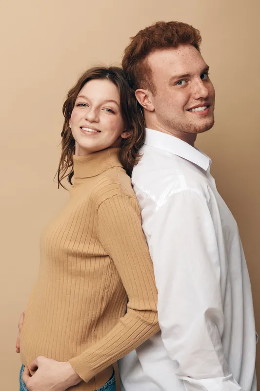 Smiling young couple stands back to back against beige background. Casual clothing, relaxed mood, and positive connection between man and woman in studio portrait.