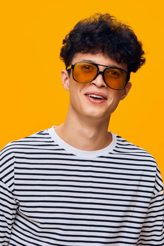 Young man wearing striped shirt and yellow sunglasses smiling confidently against vibrant orange background, showcasing cheerful and trendy style.