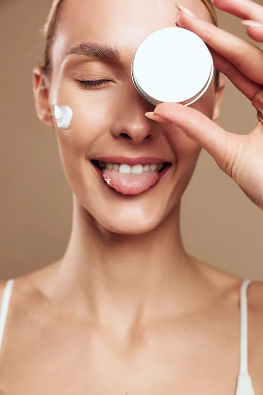 Young woman with clear skin holds a round skincare cream container near her eye while smiling playfully with her tongue out in a close-up beauty portrait on a neutral background.