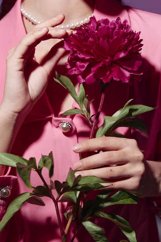 Close-up of elegant woman’s hands holding a vivid magenta peony flower with glossy leaves. She wears soft pink textured blazer and pearl necklace. Minimal beauty fashion concept.