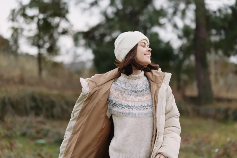 Woman in a warm coat and knit sweater standing in a park, enjoying the outdoors with a smile and wind in her hair, creating a cozy winter mood.