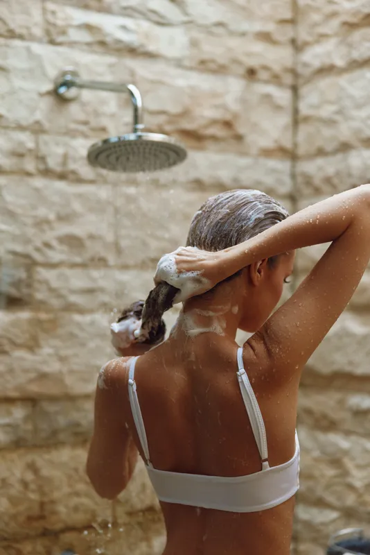 woman washing hair in outdoor shower with beige stone wall background, wet skin, white swimsuit, natural light, refreshing moment, skincare routine