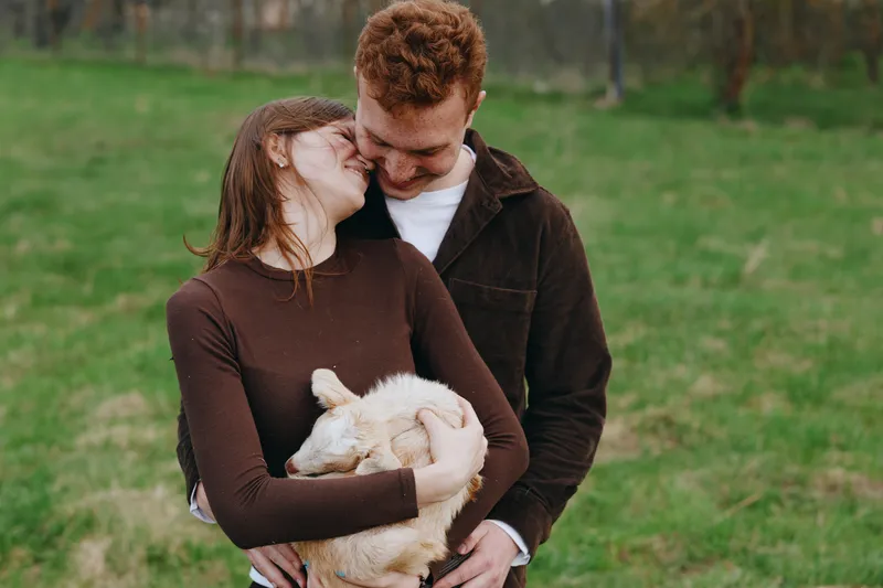 Cheerful couple outdoors, embracing a gentle goat kid in a sunlit meadow, candid affection and warmth, casual wear, natural colors, soft breeze, serene countryside mood