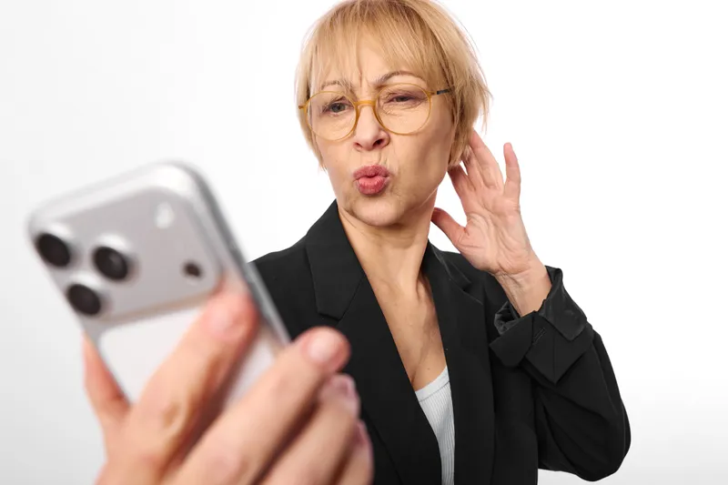 Elderly woman with glasses holding a phone with a surprised expression, studio portrait on white background, business communication concept, soft light