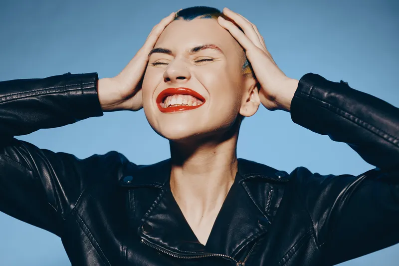 Young woman with shaved head and bright red lipstick smiling joyfully, wearing a black leather jacket against a clear blue sky background, showing excitement and freedom.
