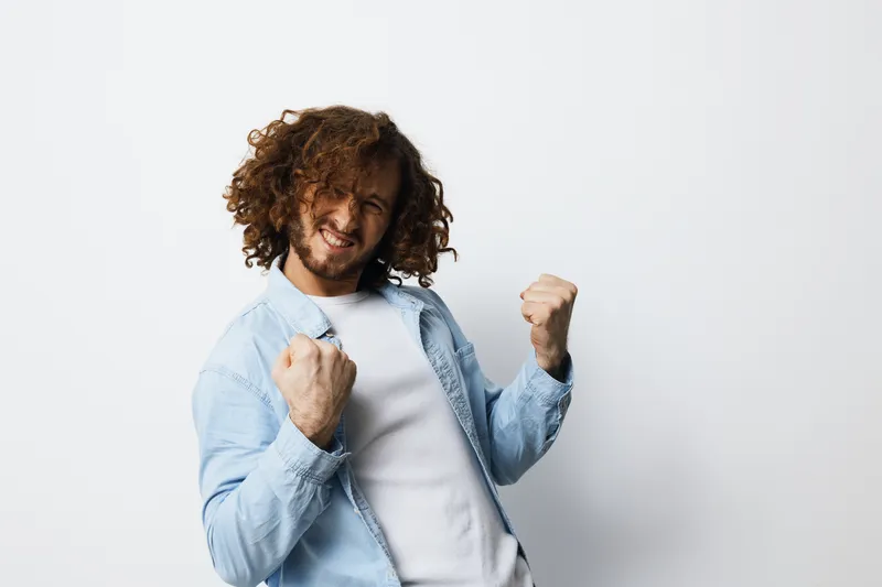 A joyful young man with curly hair celebrates success with clenched fists against a minimalistic white background, expressing confidence and happiness