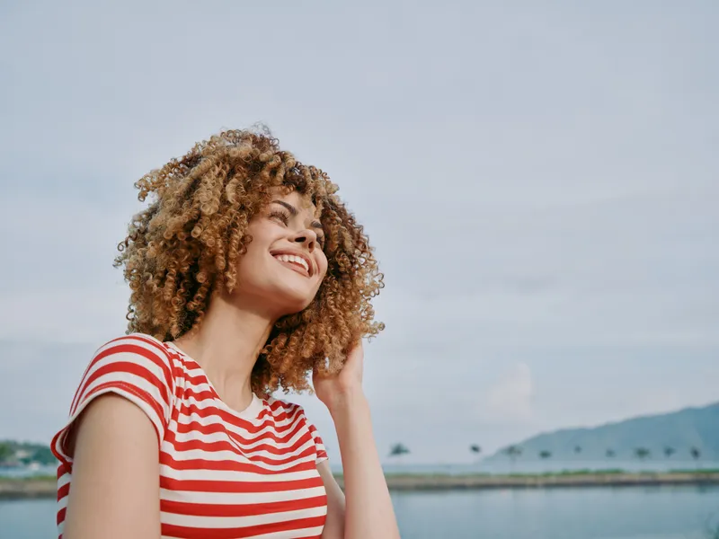 A smiling woman with voluminous curly hair wears a red and white striped shirt, gazing upward with a joyful expression against a calm lakeside backdrop and clear sky.