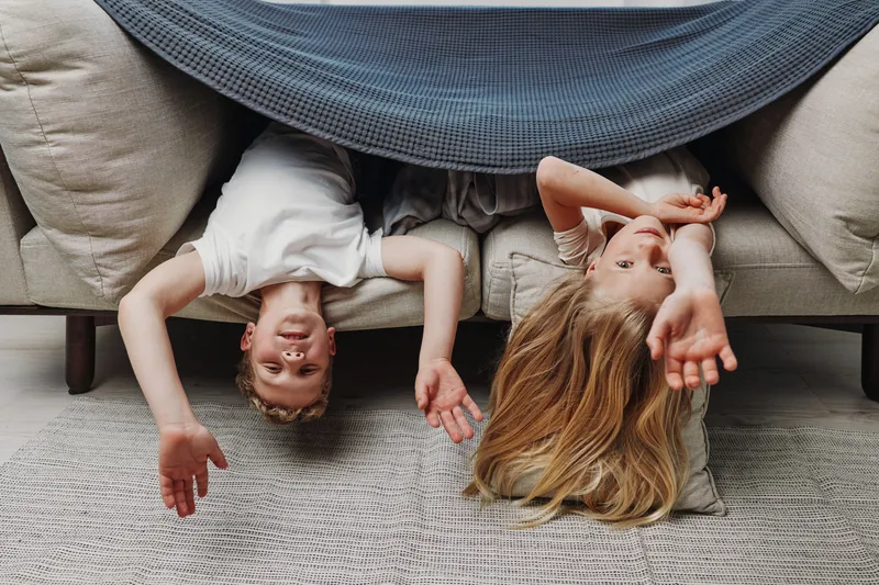 Two children lying upside down on a sofa covered with a blanket, playing and resting together in a cozy living room environment. Concept of family, childhood, and home comfort.