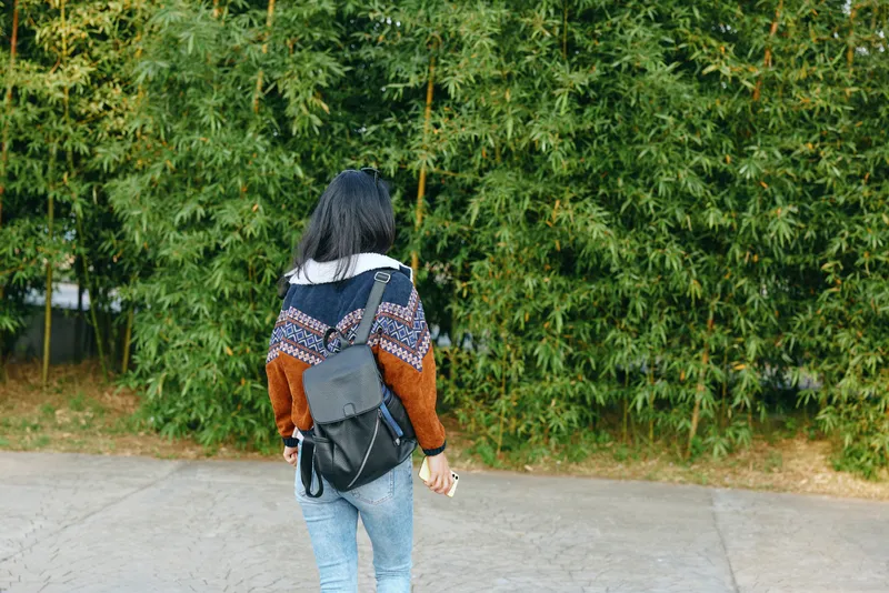 Back view of a woman wearing a patterned knit sweater and backpack, strolling along a paved path beside a dense green hedge, conveying a casual outdoor commute and relaxed travel vibe