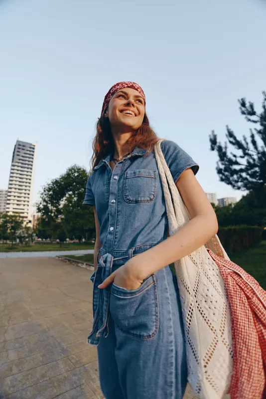 smiling woman in denim dress outdoors with shoulder bag and red headband enjoying sunny day in urban park environment