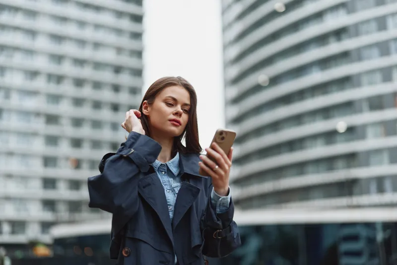 Young woman in a dark trench coat uses a smartphone outdoors in an urban environment with modern glass buildings in the background.