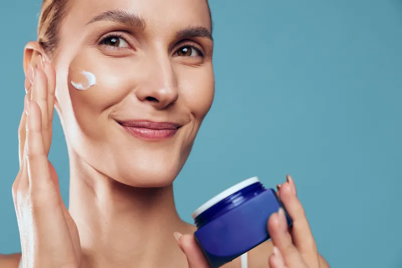 Young woman with brunette hair applies moisturizing cream on her cheek while holding a blue jar against a plain blue background, smiling gently with a relaxed expression.