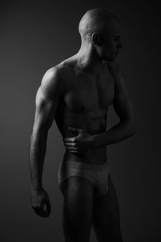 Black and white image of a muscular man posing elegantly, showcasing his strength and physique with a dramatic lighting effect for striking contrast.