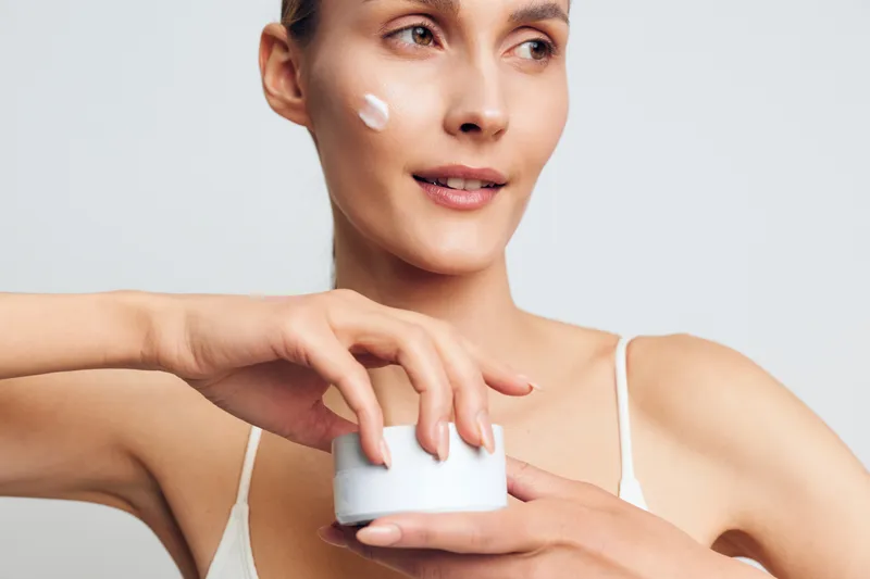 A young woman in a white tank top applies facial cream on her cheek while holding an open jar. The bright background highlights skincare and beauty routine concepts.