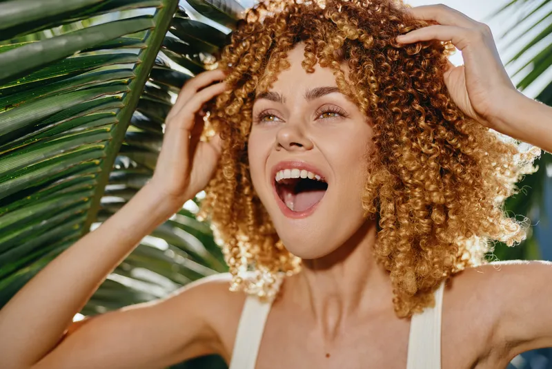 Smiling woman with curly hair enjoying sunny tropical day outdoors, expressing happiness and vitality with lush green palm leaves in background.