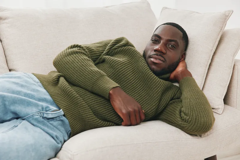 Relaxed young African man in a cozy green sweater lounging on a light colored sofa, exuding calm and comfort, perfect for lifestyle and home decor