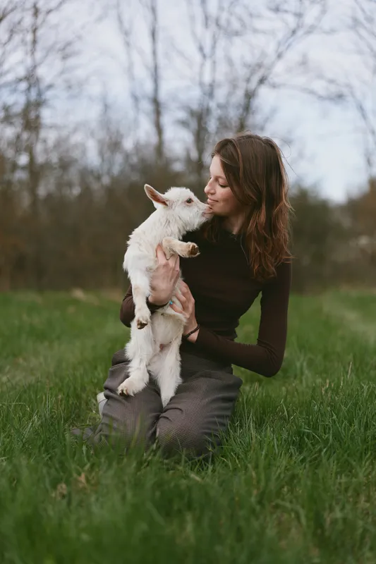 A candid lifestyle scene shows a woman outdoors, embracing a playful goat kid in a grassy field, conveying affectionate connection with animals in a calm, sunlit setting.