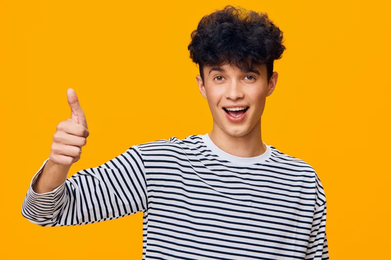 Happy young man with curly hair wearing a striped shirt shows thumbs up gesture against bright orange background, expressing positivity and enthusiasm with smile.
