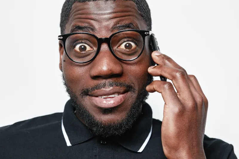 African American man with dark skin and glasses talking on smartphone with surprised expression. Closeup portrait of black male in casual black polo shirt on white background.