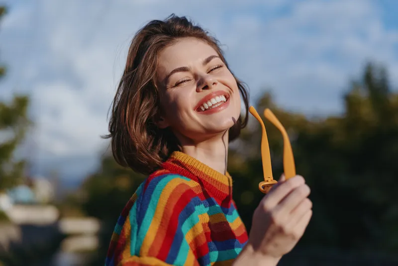 Woman smiling in a rainbow striped sweater holding yellow sunglasses outdoors, young adult female mid 20s portrait with short hair, joyful expression, casual outfit and natural sunlight lifestyle