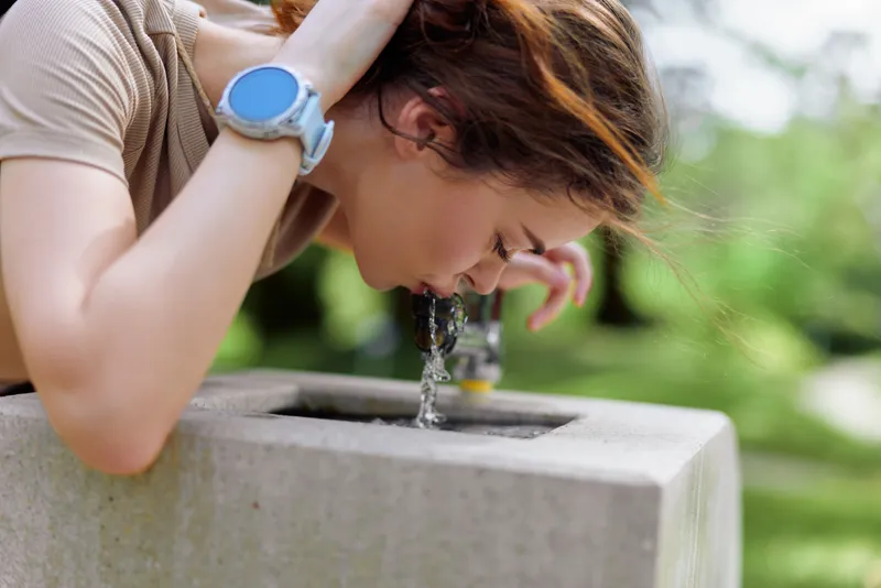 Woman drinking water from outdoor fountain in park, leaning over concrete basin for refreshment and hydration, casual portrait with smartwatch and natural green background.