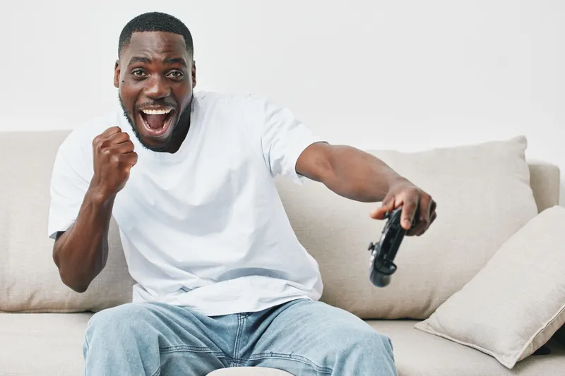 Excited young man playing video games at home on a comfortable couch, celebrating a win, showcasing joy and enthusiasm