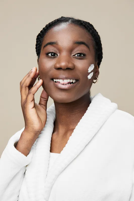 Confident young woman applying cream on her face, showcasing skincare routine with a bright smile, minimalist backdrop, and soft color palette