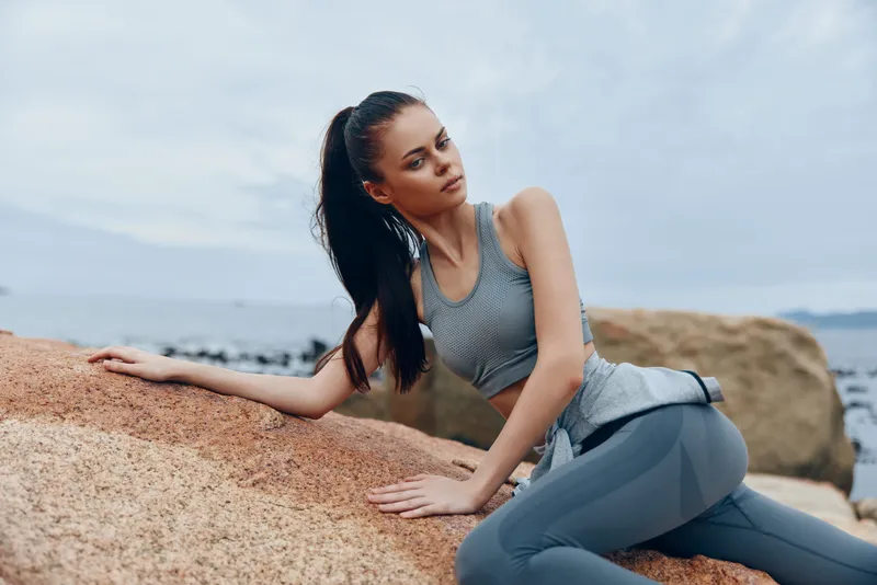 Young athletic female model in activewear posing on a rocky beach, embodying fitness and confidence against a serene ocean backdrop