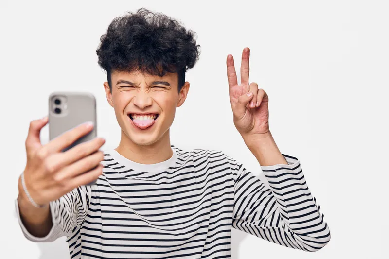 young man taking selfie with smartphone, playful expression, striped shirt, curly dark hair, isolated white background, casual style, fun gesture