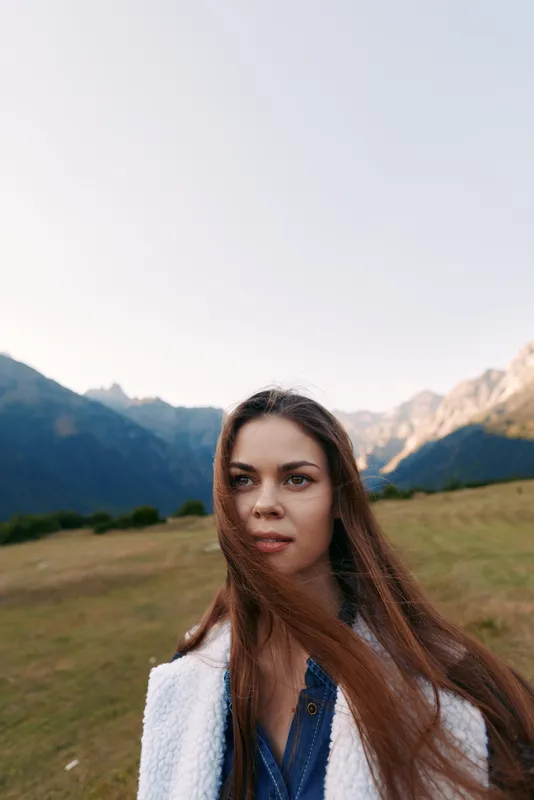 Woman portrait in mountains meadow with longhair blowing in wind, nature outdoor scene, casual young female looking thoughtful, scenic landscape and soft natural light