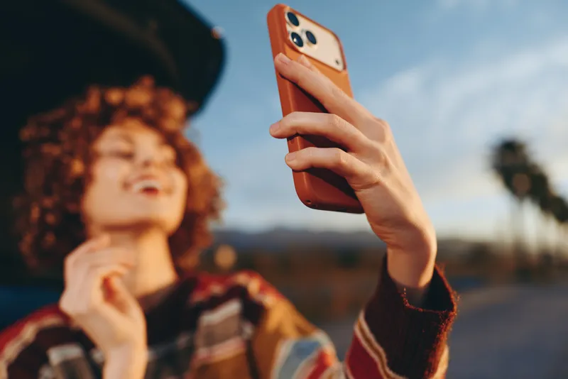 Woman holding smartphone for selfie outdoors, street travel lifestyle mood, curly hair woman in background, blurred phone hands, soft daylight, bokeh street landscape, casual fashion