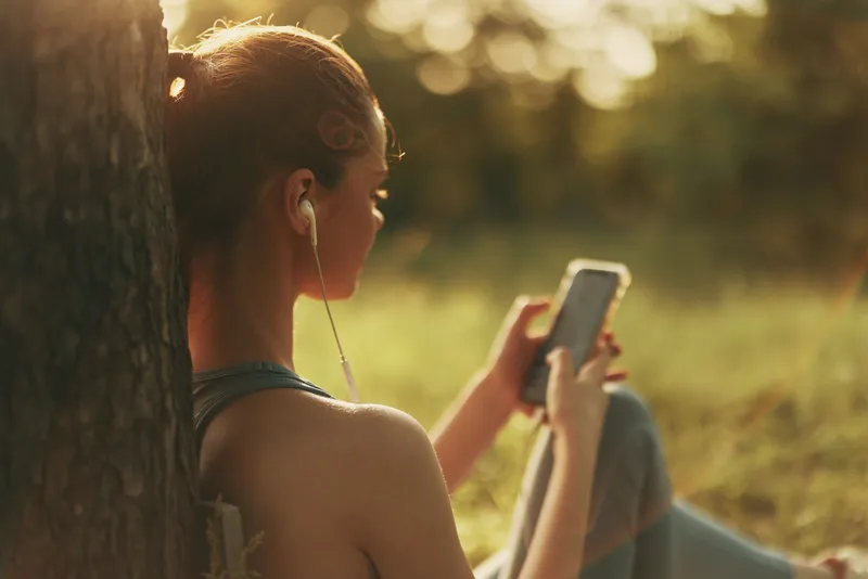 Young woman enjoying music on smartphone outdoors, sitting against a tree in natural sunlight, capturing relaxation and tranquility in a vibrant green setting