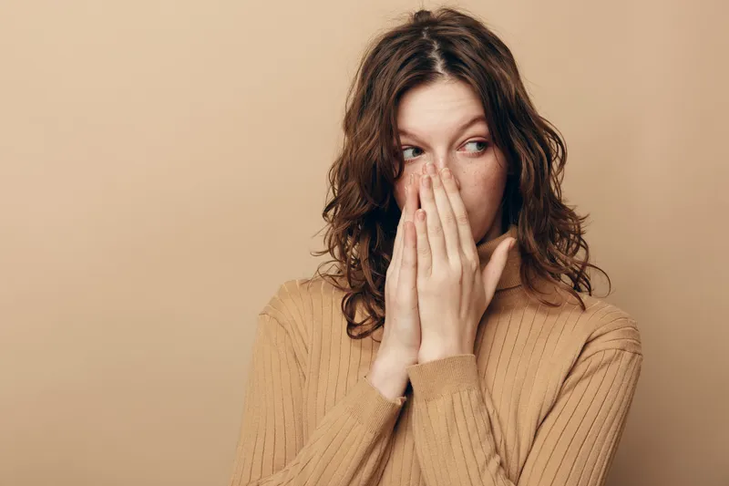 Young woman with curly hair wearing beige sweater covers mouth with hands looking sideways. Neutral background and soft lighting create a thoughtful and introspective mood.