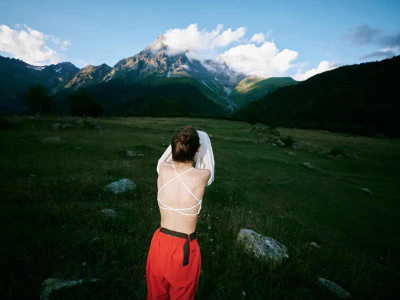 Back view of a figure in red pants and white strappy top standing in a green meadow, facing towering mountains under blue sky with fluffy clouds, capturing remote outdoor adventure.