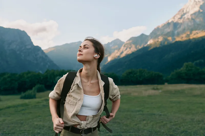 Young woman with backpack enjoys mountain landscape during hiking trip, wearing casual outfit and wireless earbuds, expressing calm and confident lifestyle outdoors.
