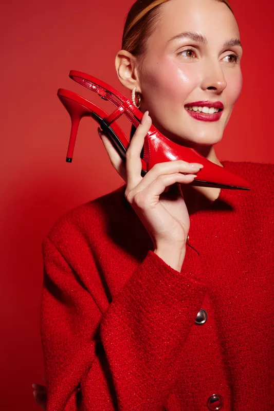 Smiling woman in red sweater holds red high heel shoe near face on red background. Fashion concept with bright colors, glamour style, and confident female model posing in studio lighting.