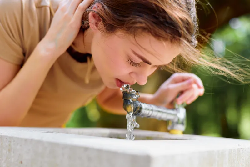 Woman drinking water from a public fountain outdoor in a sunlit park, summer portrait closeup capturing hair movement, natural skin detail and a refreshing candid moment of hydration.