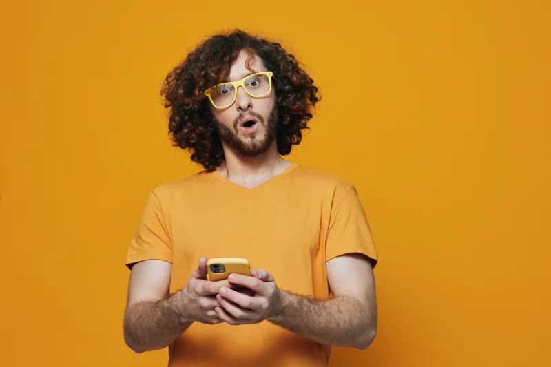 Surprised young man with curly hair in bright yellow glasses, holding a smartphone against a vibrant yellow background, showcasing emotion and modern lifestyle