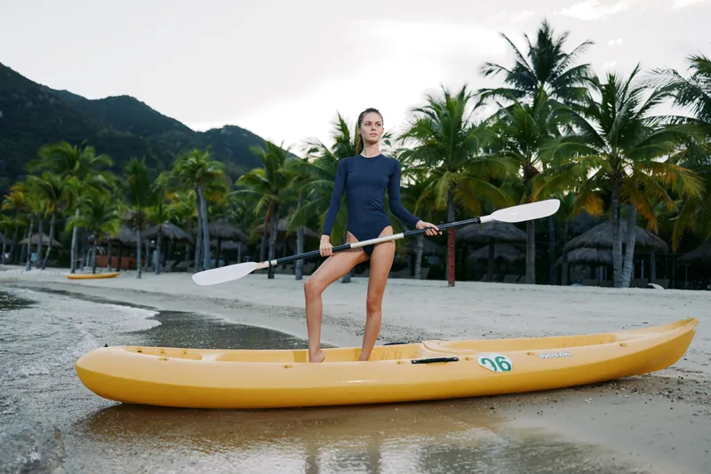 A confident woman stands on a bright yellow kayak at the shore, paddles in hand, ready for adventure as surf and palm trees frame a tropical coastline.