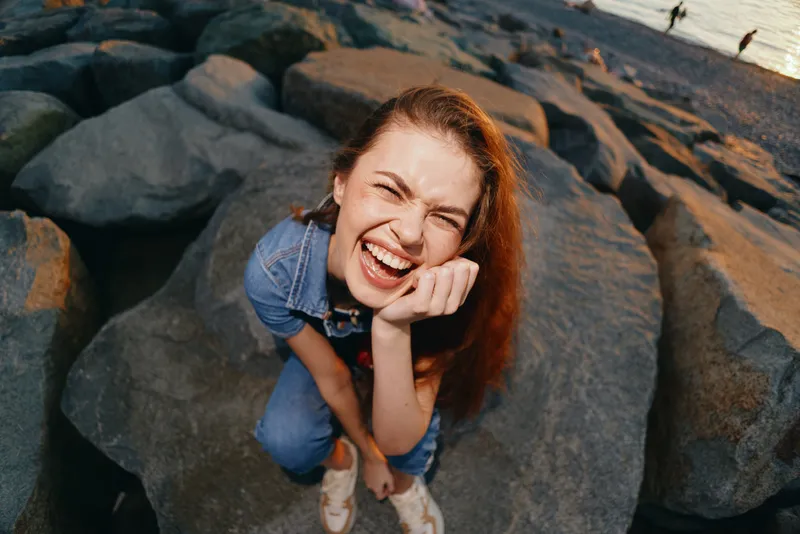 lifestyle woman in modern boho-western denim street style laughing joyfully on rocky beach with warm film color tones, capturing natural happiness and casual fashion