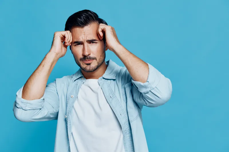 Handsome man with a serious expression holds his head with hands, showcasing a moment of stress or concern against a vibrant blue background. Emotion concept