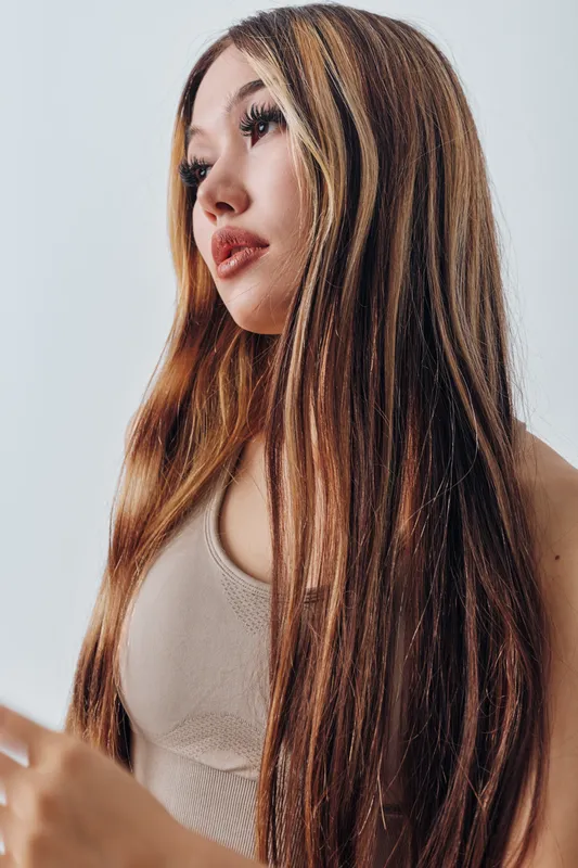 Young woman with long straight hair and natural makeup wearing a sleeveless beige top looking sideways on white background. Beauty portrait and casual style with confident expression.